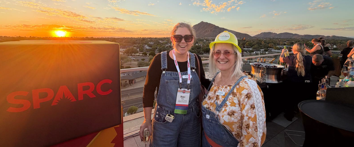 Staci and Nomi smile beside a SPARC sign on a terrace, with mountains and a golden sunset behind them.